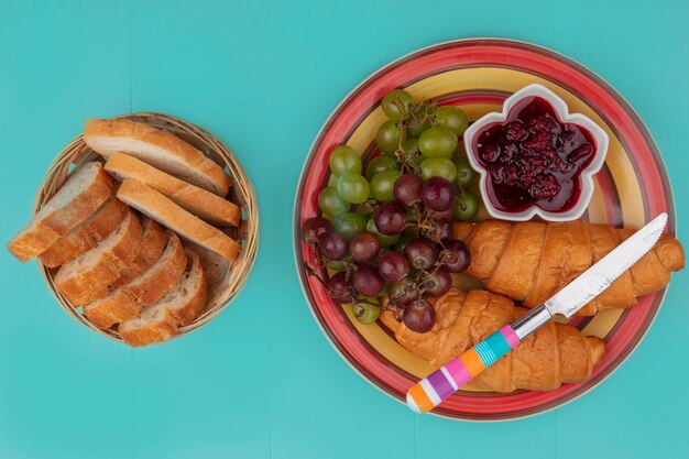 Vista superior do café da manhã com croissant, geleia de uva e framboesa e fatias de pão com faca sobre fundo azul