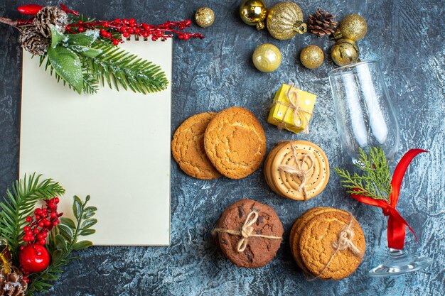 Vista superior do cálice de vidro caído com fita vermelha e acessórios de decoração ao lado do caderno de biscoitos empilhados em fundo escuro