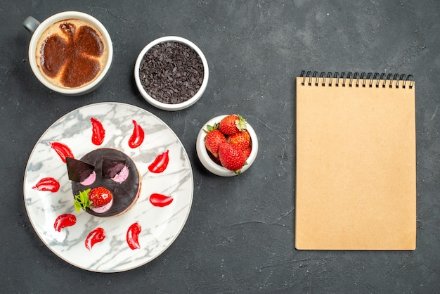 Vista superior do cheesecake de morango em uma tigela oval branca com morangos e chocolate uma xícara de café um caderno na superfície escura