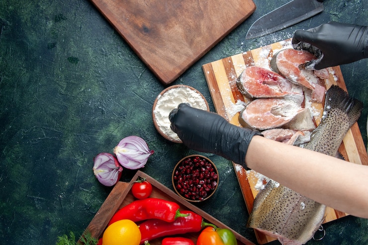 Foto grátis vista superior do chef cobrindo fatias de peixe cru com farinha de legumes frescos na tábua de madeira faca tigela de farinha na mesa da cozinha