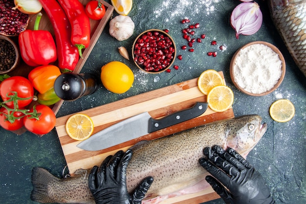 Vista superior do chef com luvas pretas segurando peixe cru na tábua de madeira Moedor de pimenta farinha tigela sementes de romã na tigela na mesa da cozinha