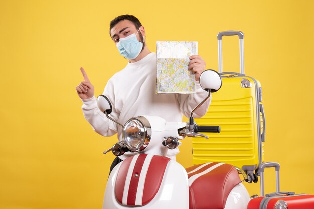 Vista superior do conceito de viagem com um cara confiante em uma máscara médica em pé perto da motocicleta com uma mala amarela e segurando o mapa apontando para cima