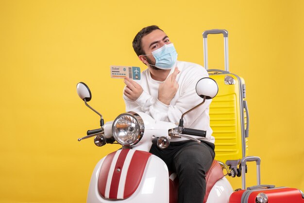 Vista superior do conceito de viagem com um cara esperançoso com uma máscara médica sentado na motocicleta com uma mala amarela e segurando a passagem