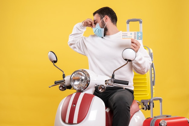 Vista superior do conceito de viagem com um jovem com máscara médica sentado em uma motocicleta com uma mala amarela e segurando a passagem, fazendo um gesto de mau cheiro