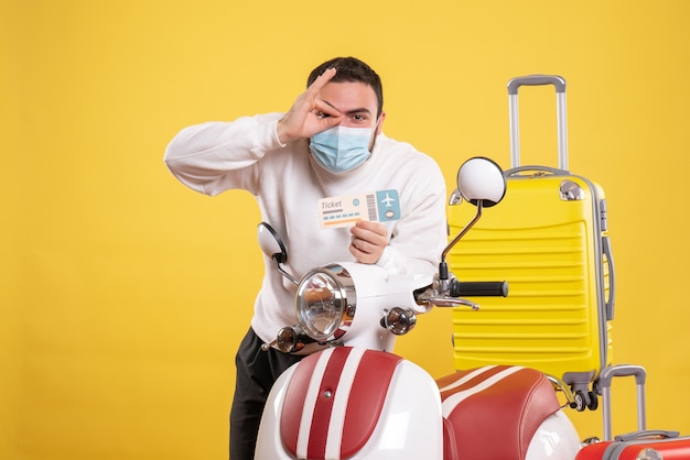 Vista superior do conceito de viagens com um cara confiante com uma máscara médica em pé perto da motocicleta com uma mala amarela e segurando o bilhete