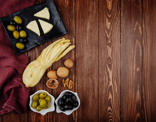 Vista superior do cream cheese em forma de triângulo com azeitonas em conserva em uma bandeja preta e nozes com queijo de corda na mesa de madeira escura com espaço de cópia