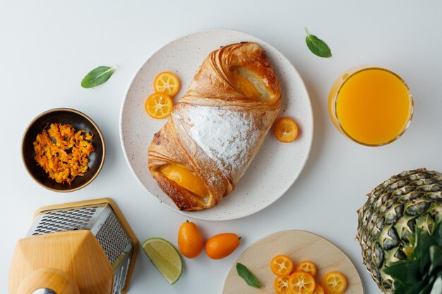 Vista superior do croissant com fatias de kumquat em prato e suco de laranja de abacaxi com ralador em fundo branco