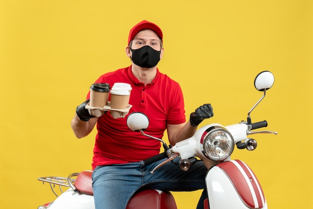 Vista superior do entregador ambicioso usando uniforme e luvas de chapéu, máscara médica, sentado na scooter, mostrando pedidos