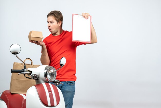 Vista superior do entregador surpreso de uniforme vermelho em pé perto da scooter, segurando o pedido e o documento na parede branca