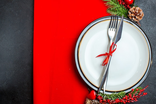 Vista superior do fundo de Natal com talheres com fita em um prato de jantar acessórios de decoração ramos de pinheiro em um guardanapo vermelho no lado esquerdo em um fundo escuro