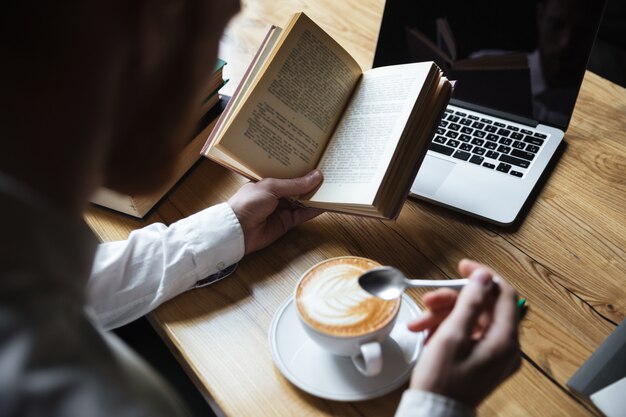 Vista superior do homem de camisa branca, mexendo o café durante a leitura do livro