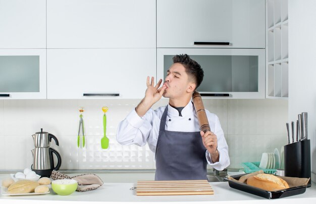Vista superior do jovem cozinheiro de uniforme em pé atrás da mesa, segurando o ralador e fazendo um gesto perfeito na cozinha branca