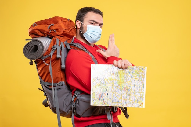 Vista superior do jovem viajante que pensa, usando máscara médica com uma mochila segurando um mapa apontando para cima em amarelo