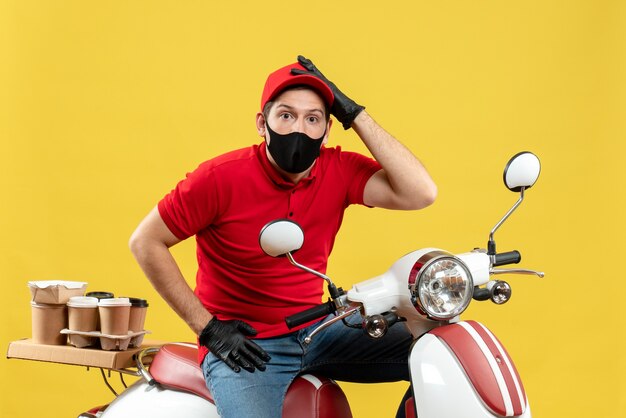 Vista superior do mensageiro confuso usando blusa vermelha e luvas de chapéu com máscara médica, entregando pedidos sentado na scooter