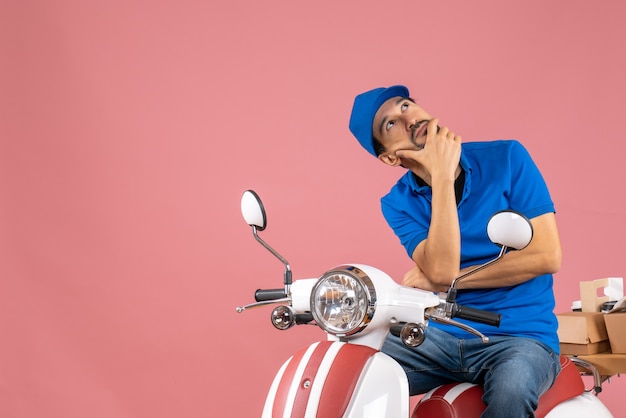 Foto grátis vista superior do mensageiro sonhador usando um chapéu, sentado na scooter, entregando pedidos em fundo cor de pêssego