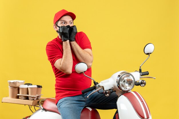 Vista superior do mensageiro usando blusa vermelha e luvas de chapéu na máscara médica, entregando pedidos sentado na scooter, olhando para algo com expressão facial de medo