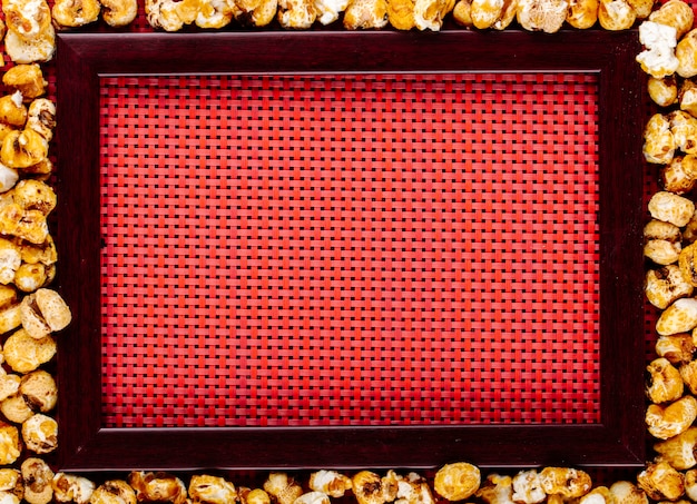 Vista superior do pipoca caramelizada doce espalhada ao redor da moldura vazia sobre fundo vermelho, com espaço de cópia