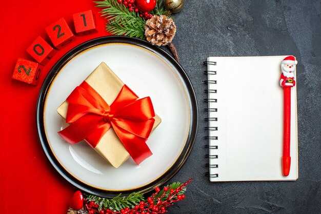 Vista superior do plano de fundo do ano novo com o presente no prato de jantar acessórios de decoração ramos de abeto e números em um guardanapo vermelho e caderno com caneta em uma mesa preta