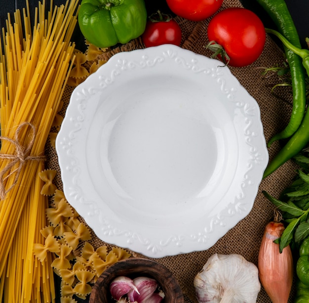 Vista superior do prato branco com macarrão cru e espaguete, tomate, alho, cebola e pimenta em um guardanapo bege