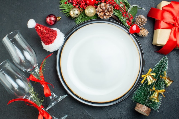 Vista superior do prato de jantar árvore de Natal ramos de abeto coníferas cone caixa de presente chapéu de Papai Noel cálices de vidro caídos em fundo preto