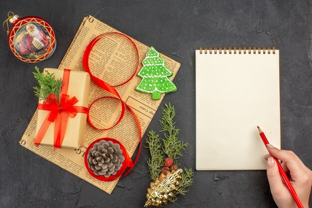 Vista superior do presente de Natal em papel pardo com fita de abeto no jornal Enfeites de Natal e lápis bloco de notas em mão feminina na superfície escura