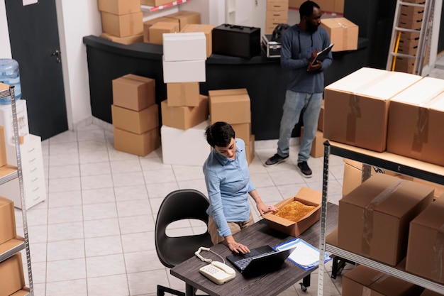 Vista superior do supervisor do armazém embalando o pedido de roupas do cliente em caixa de papelão enquanto verifica os detalhes de envio no computador portátil. Equipe trabalhando no armazém no departamento de entrega.