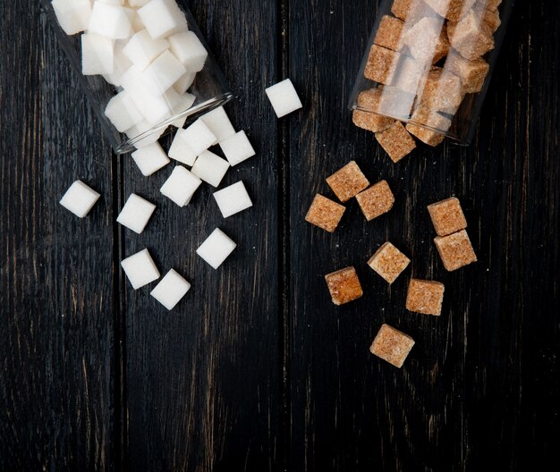 Vista superior dos cubos de açúcar branco e marrom espalhados de frascos de vidro no fundo escuro de madeira