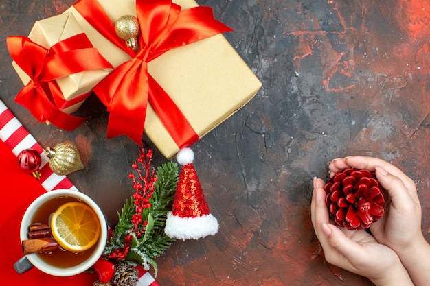 Vista superior dos presentes de Natal amarrados com uma pinha de chapéu de Papai Noel de fita vermelha em uma xícara de chá feminina na mesa vermelha escura