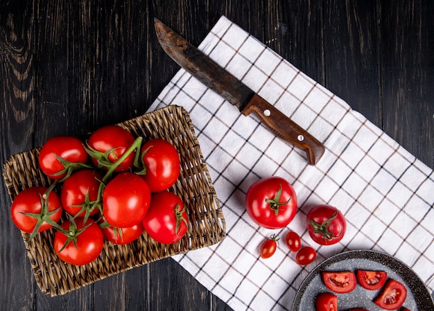 Vista superior dos tomates no prato de cesta com outros no pano e faca na superfície de madeira