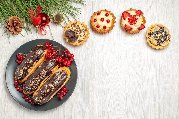 Vista superior éclairs de chocolate e groselhas nas tortas de prato cinza e folhas de pinheiro com brinquedos de natal na mesa de madeira branca