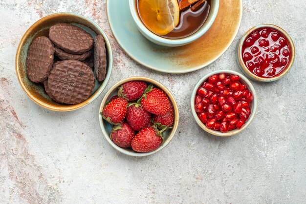 Vista superior em close-up de morangos, uma xícara de chá preto com limão e paus de canela, tigelas de diferentes frutas, biscoitos de chocolate e sementes de romã na mesa