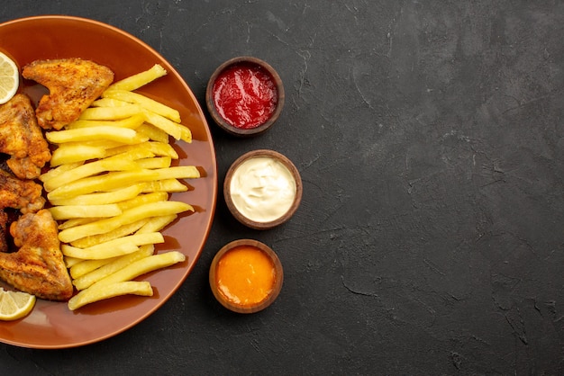 Vista superior em close-up prato de fastfood com batatas fritas, asas de frango e limão e três tigelas de diferentes tipos de molhos na mesa escura