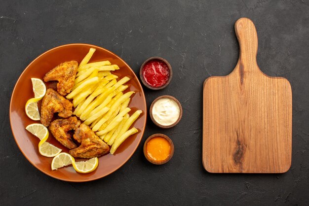 Vista superior em close-up prato fastfood de asas de frango, batata frita e limão ao lado de tigelas de três tipos de molhos e tábua de cozinha de madeira na mesa