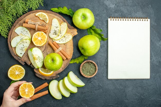 Vista superior, maçãs verdes, paus de canela e fatias de limão fatias de maçã na placa de madeira corte limão no caderno de maçãs de mão feminina na mesa preta