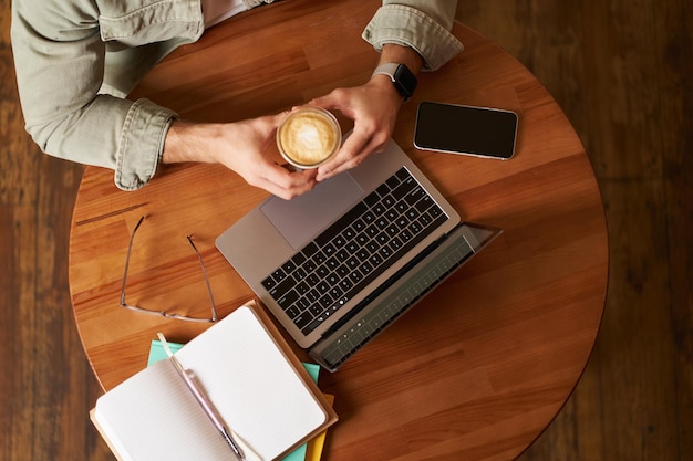 Vista superior mãos masculinas segurando uma xícara de café homem sentado em uma mesa redonda em um café trabalhando em um laptop tinha