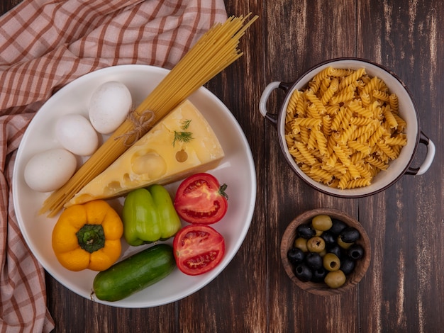 Vista superior queijo maasdam com frango ovos tomate pepino espaguete cru e pimentão em um prato e macarrão cru em uma panela sobre um fundo de madeira