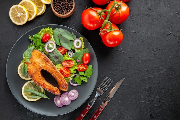 Vista superior saboroso peixe cozido com legumes frescos e talheres em uma mesa escura