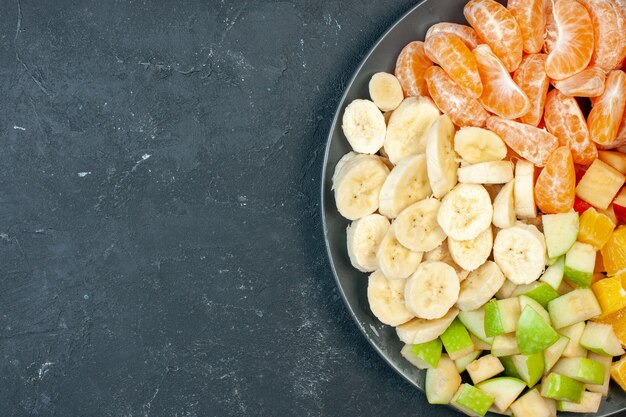 Vista superior salada de frutas frescas fatiadas bananas, maçãs e laranjas em fundo escuro