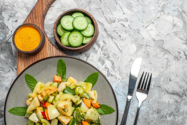 Vista superior salada de tomate verde em prato oval açafrão na tigela de tábua com corte garfo e faca de pepino em fundo escuro