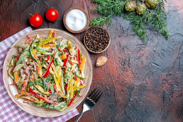 Vista superior salada de vegetais no prato na toalha de mesa garfo sal e pimenta preta tomate ramos de pinheiro no espaço livre da mesa vermelha escura