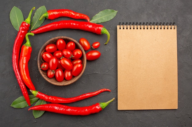 Vista superior semicírculo de pimentas vermelhas e folhas de louro e uma tigela de tomates cereja e um caderno na mesa preta