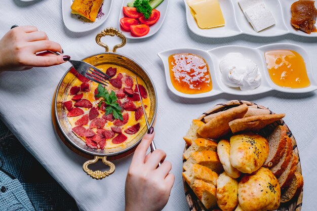 Vista superior uma mulher tomando omelete de café da manhã com salsicha em uma panela com uma cesta de pão