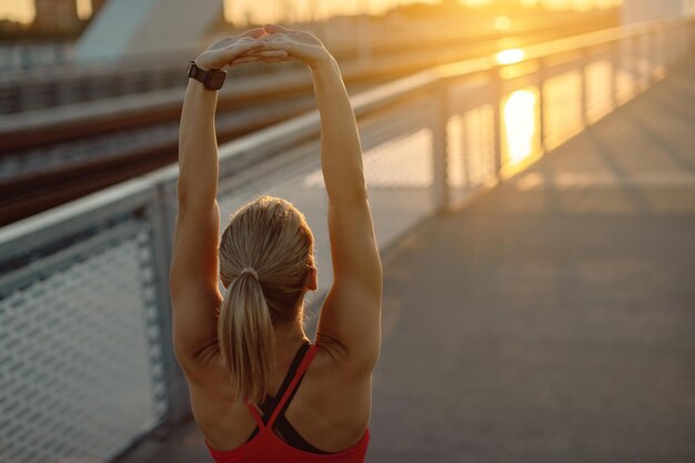 Vista traseira da atleta feminina, estendendo-se em uma ponte ao pôr do sol