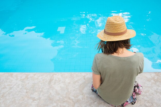Foto grátis vista traseira da mulher de chapéu de palha verão sentado à beira da piscina