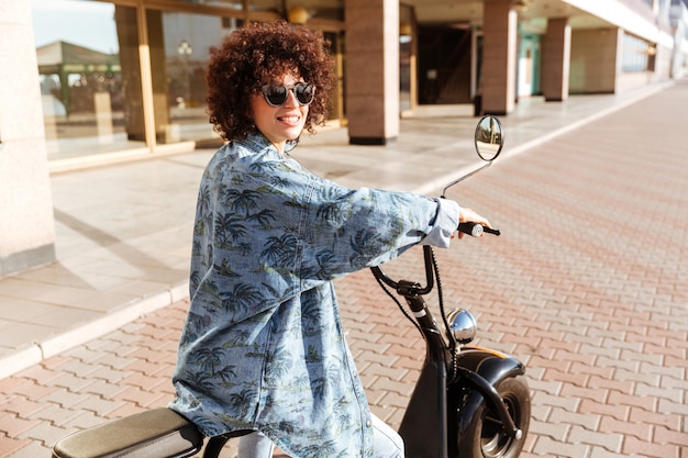 Vista traseira da mulher encaracolada feliz em sunglasess sentado na moto moderna ao ar livre e desviar o olhar