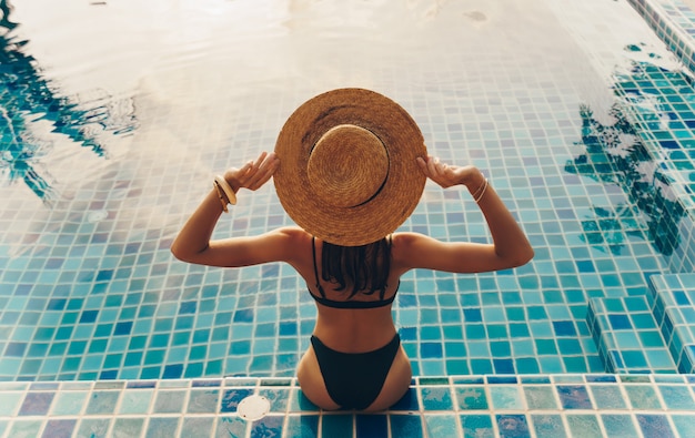 Vista traseira da mulher graciosa em traje de banho e chapéu, sentado perto da piscina