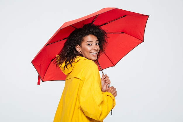 Vista traseira da mulher sorridente na capa de chuva se escondendo sob o guarda-chuva