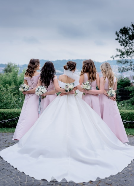 Vista traseira da noiva em vestido de noiva e damas de honra vestidas nos vestidos-de-rosa iguais ao ar livre