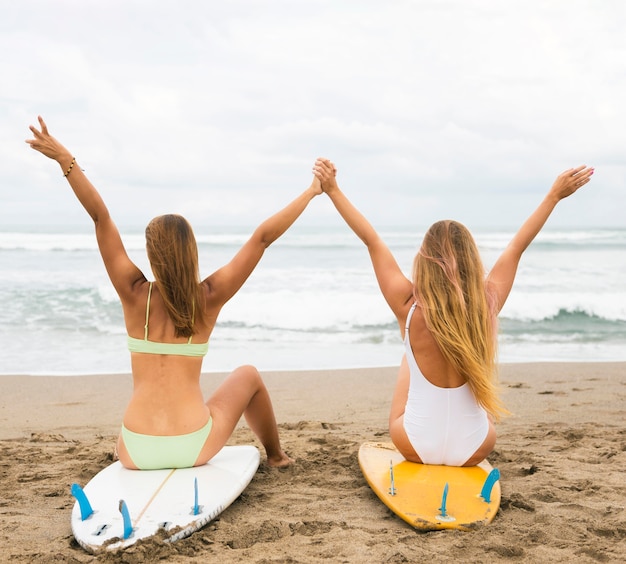 Vista traseira de amigas na praia em pé na prancha de surf com as mãos para cima