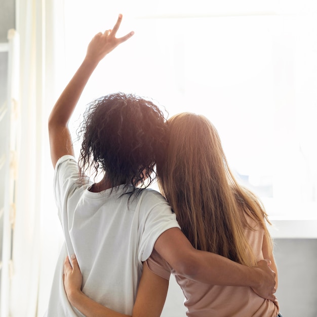 Vista traseira de duas amigas se abraçando em frente à janela